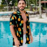 pexels-photo-3297861-3297861 A young man smiling by the pool, wearing a colorful pineapple shirt and white shorts.