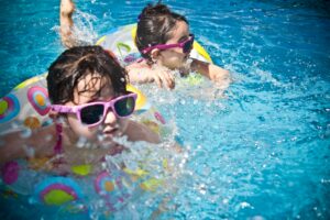 Two young girls enjoying a playful day in a bright blue swimming pool with colorful float rings, Pool care in Houston TX by Memorial Pool Services