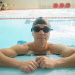 pexels-photo-6539736-6539736 Adult male swimmer wearing goggles and cap relaxing at the edge of an indoor pool.